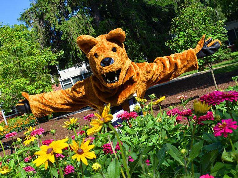 The Nittany Lion holding out his arms in a welcoming way behind a bed of flowers.